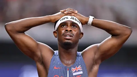 Getty Images Jeremiah has his hands on his head and is looking up. He is wearing a white "100% Jesus" sweat band on his forehead. He is wearing a GB vest with dark blue and white stripes. 