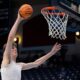 Olivier Rioux warming up before a game. He has dark hair and is wearing a white sports vest. He is holding a ball in his raised right hand as he approaches the basket to score.