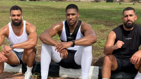 Social Media Latif Ayodele sits with Anthony Joshua and Sina Ghami by grass outside, all in training gear