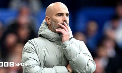 Enzo Maresca, looks on during the Premier League match between Chelsea and Brighton at Stamford Bridge. He is wearing a grey coat and holding his face.