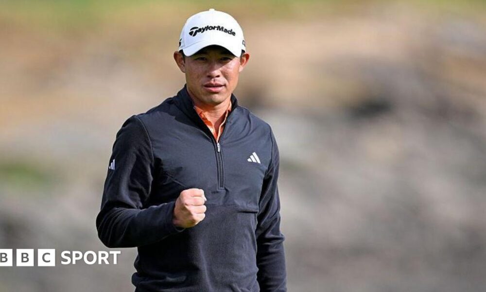 Collin Morikawa clenches his right fist after sinking his winning putt. He is wearing a white cap and black zipped-up top.