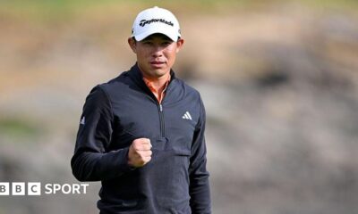 Collin Morikawa clenches his right fist after sinking his winning putt. He is wearing a white cap and black zipped-up top.