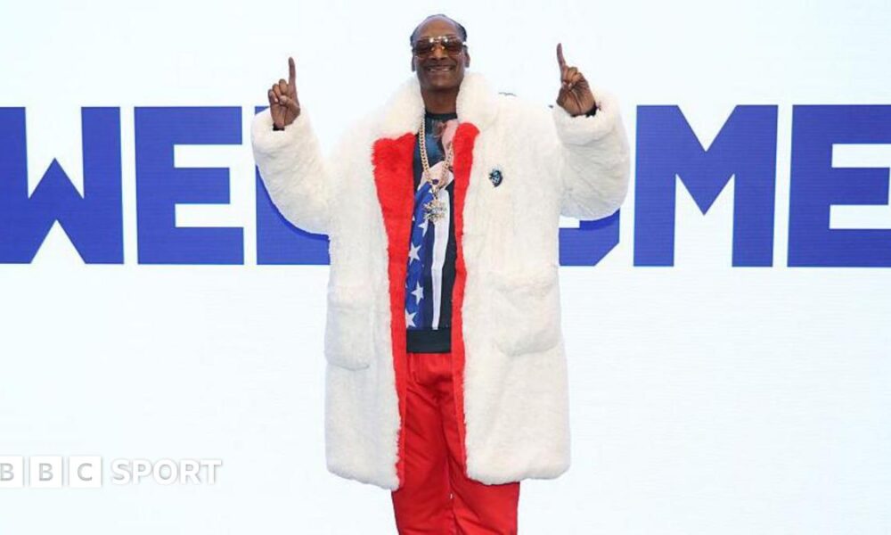 Snoop Dogg wears a fur coat over a red tracksuit, fingers pointed upwards while standing in front of a large sign saying 'Welcome' at the Winter Olympics