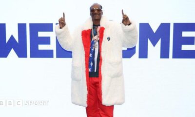 Snoop Dogg wears a fur coat over a red tracksuit, fingers pointed upwards while standing in front of a large sign saying 'Welcome' at the Winter Olympics