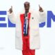 Snoop Dogg wears a fur coat over a red tracksuit, fingers pointed upwards while standing in front of a large sign saying 'Welcome' at the Winter Olympics