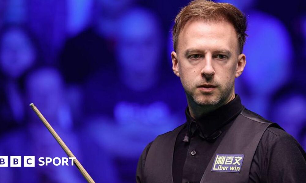 Judd Trump in black shirt and waistcoat carrying a snooker cue