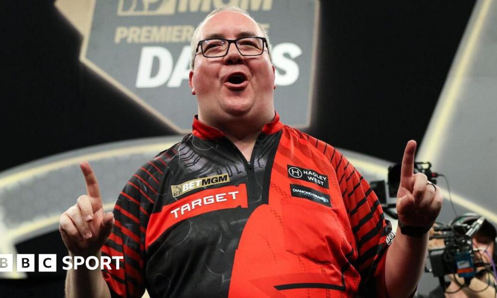 Stephen Bunting celebrates, pointing with both index fingers and pulling a celebratory facial pose as he wears a black and red darts shirt