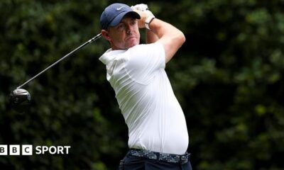 Rory McIlroy at the Truist Championship, wearing a white golf shirt and blue cap, swinging a metal wood