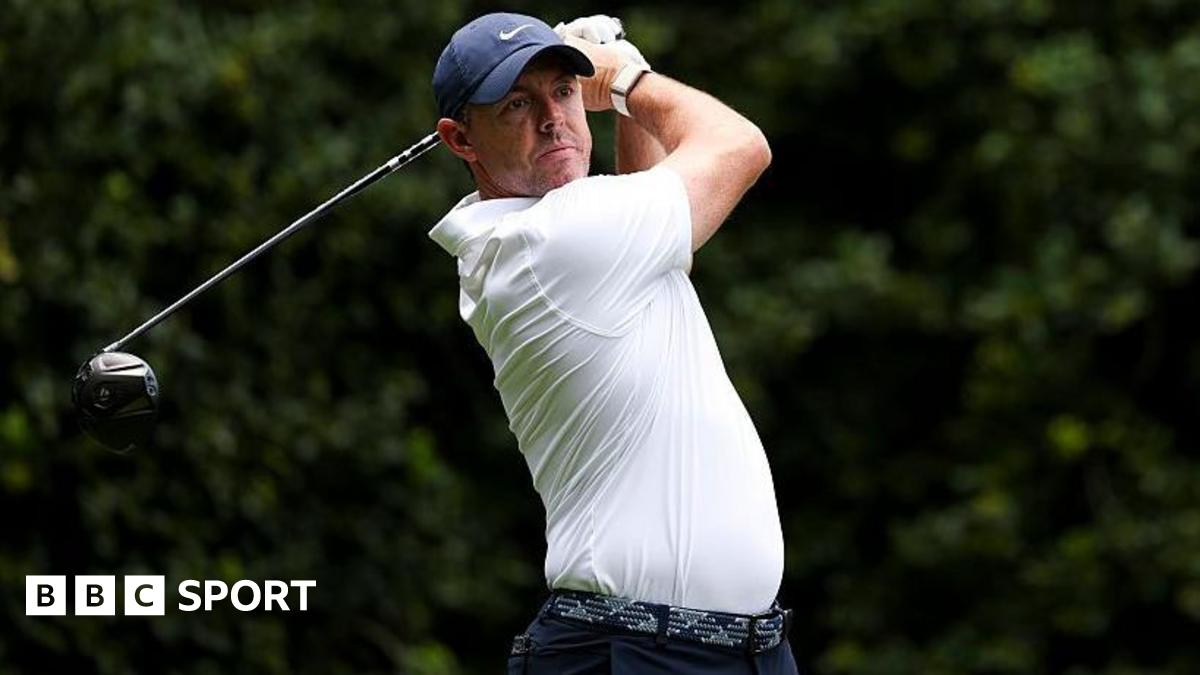 Rory McIlroy at the Truist Championship, wearing a white golf shirt and blue cap, swinging a metal wood