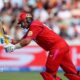 Joe Root, wearing red trousers, shirt and helmet, leans forward to play a shot with his bat held up in front of him