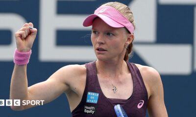 Harriet Dart, wearing a purple dress and pink visor, holds her racquet in her left hand and raises a clenched right fist