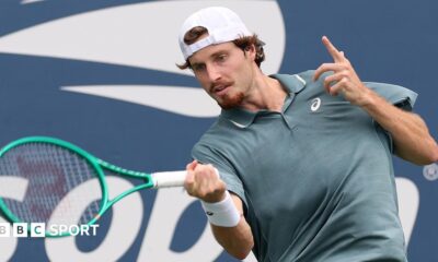 Toby Samuel, wearing a grey shirt and white backwards baseball cap, hits the ball with a green racquet